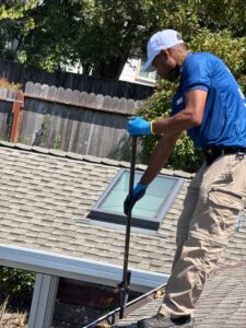 A Spotfree Window technician using a pole tool to clean gutters near a skylight on a steep, asphalt shingled residential roof.