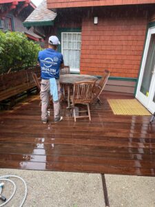 A Spotfree Window technician in a blue uniform standing on a wet, clean wooden deck during a rain shower after performing power washing services.