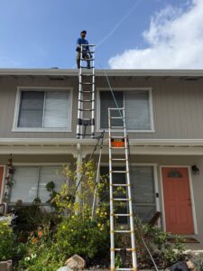 A Spotfree Window technician wearing a blue uniform, standing securely at the top of a large extension ladder to clean gutters on a two-story residential home.