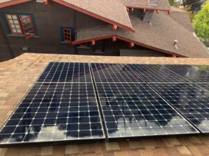 A detailed shot of dark solar panels on a brown roof, perfectly clean and wet after a professional wash by Spotfree Window, reflecting a red-trimmed house and blue sky.