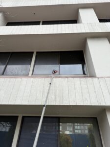 A commercial window cleaning technician using a water-fed pole system to safely clean a high-up window on a multi-story building from the ground level.